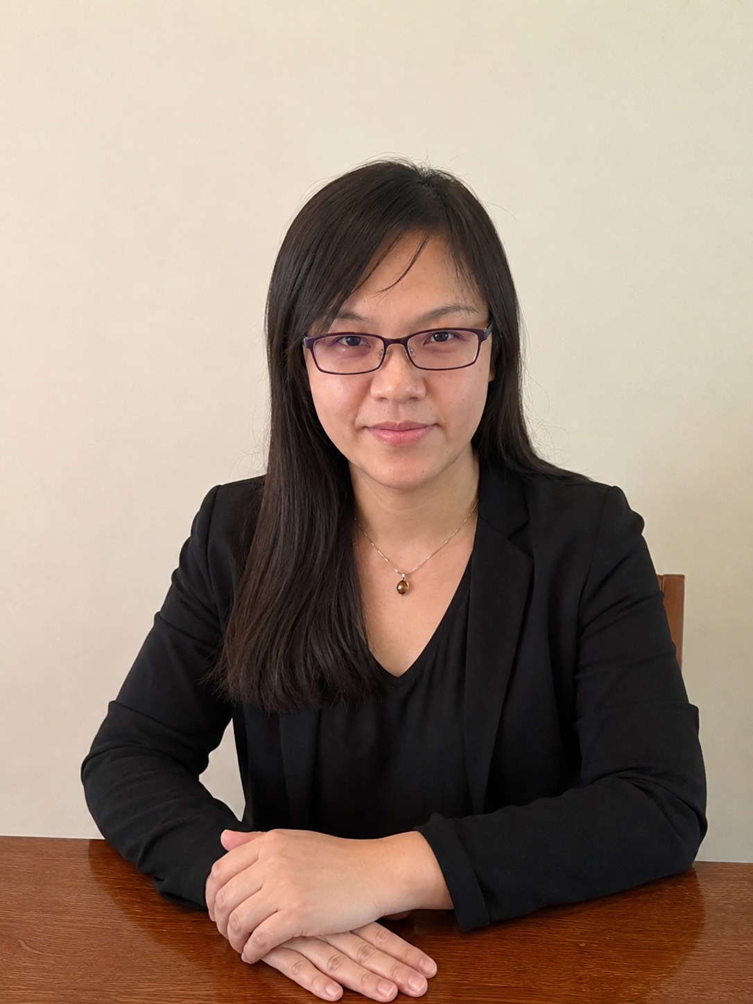 戴黑框眼鏡的女性正面半身照，穿黑色西裝外套，雙手交疊坐在木桌前。