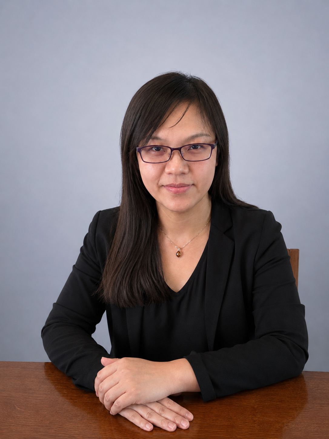 戴黑框眼鏡的女性正面半身照,穿黑色西裝外套,雙手交疊坐在木桌前。